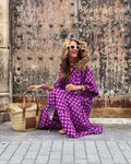 vrouw in zomerse dames vintage paarse bloemenjurk zittend met zonnebril en strooien tas tegen oude metalen deur