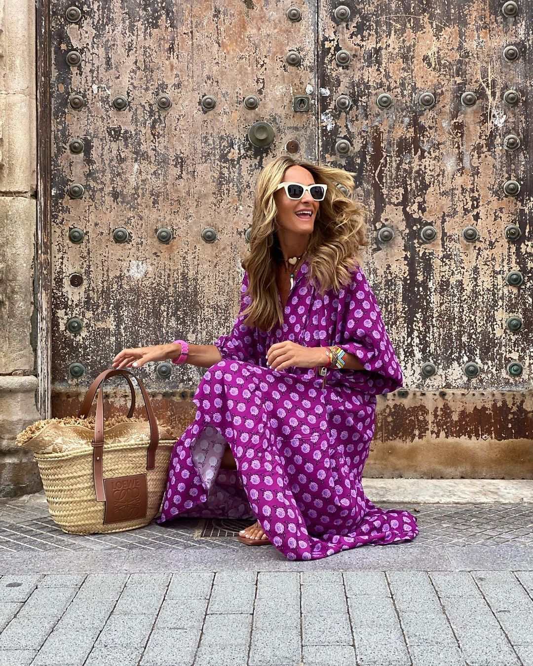 vrouw in zomerse dames vintage paarse bloemenjurk zittend met zonnebril en strooien tas tegen oude metalen deur
